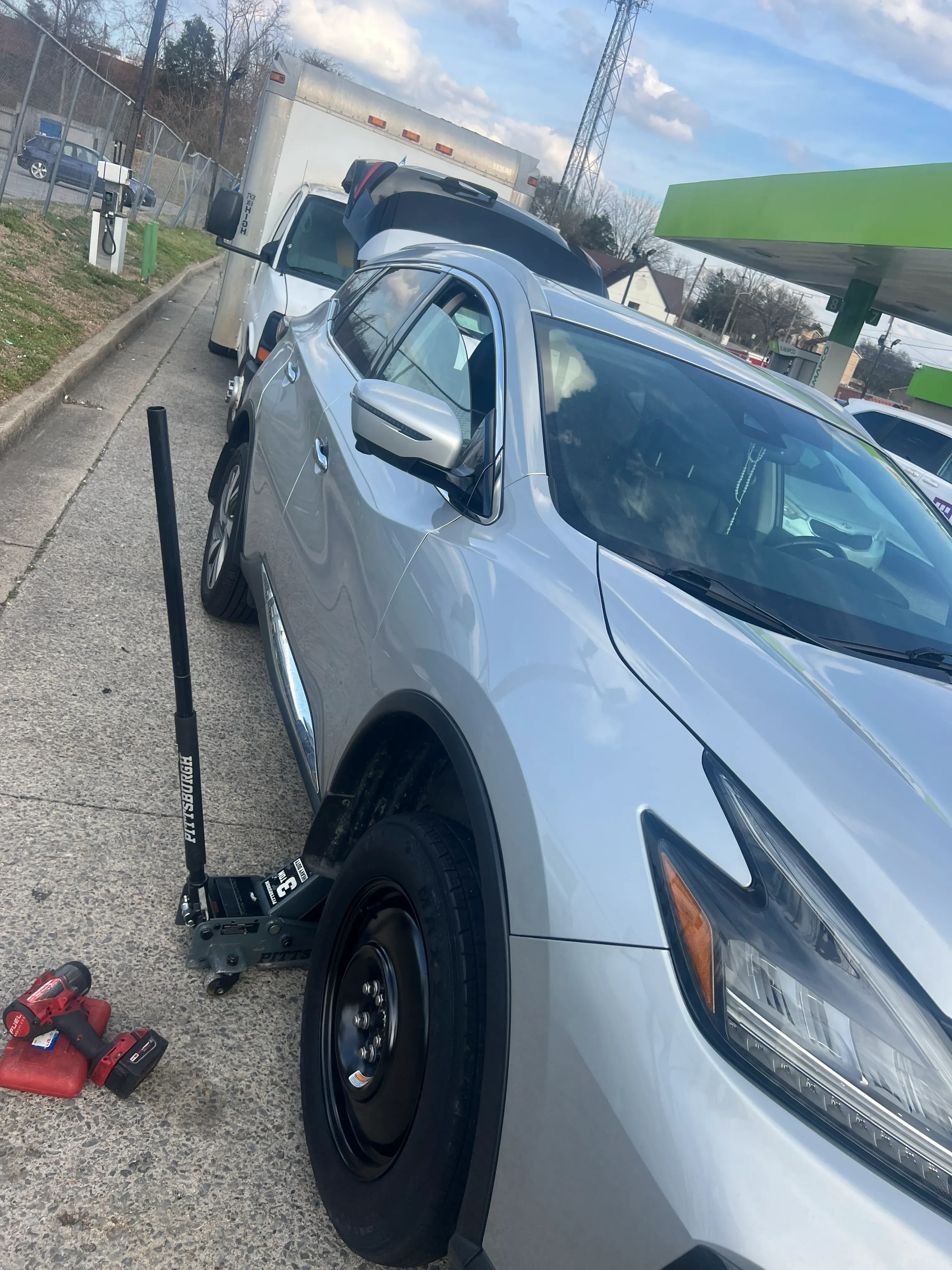 Nashville Flat Tire Service technician changing a flat tire on a silver Nissan Murano SUV at a Greater Nashville gas station, with the front-right wheel lifted on a hydraulic floor jack.